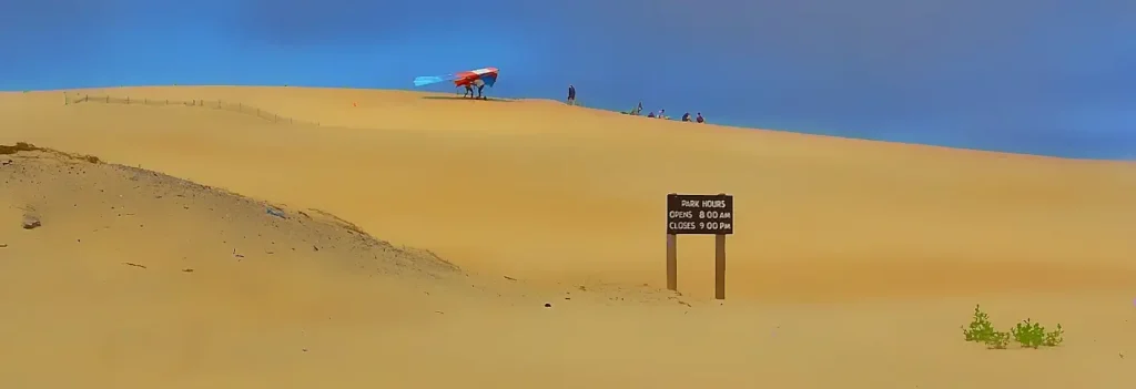 Golden sand dunes at Jockey's Ridge State Park with a park hours sign in the foreground and people with a colorful hang glider preparing on the dune ridge under a blue sky.