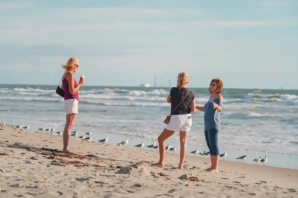 Three women standing barefoot on the beach, chatting and enjoying each other’s company near the water’s edge, with seagulls scattered on the sand and gentle ocean waves behind them under a bright sky.