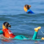 Surf contest competitors in colored jerseys paddling out through the swell
