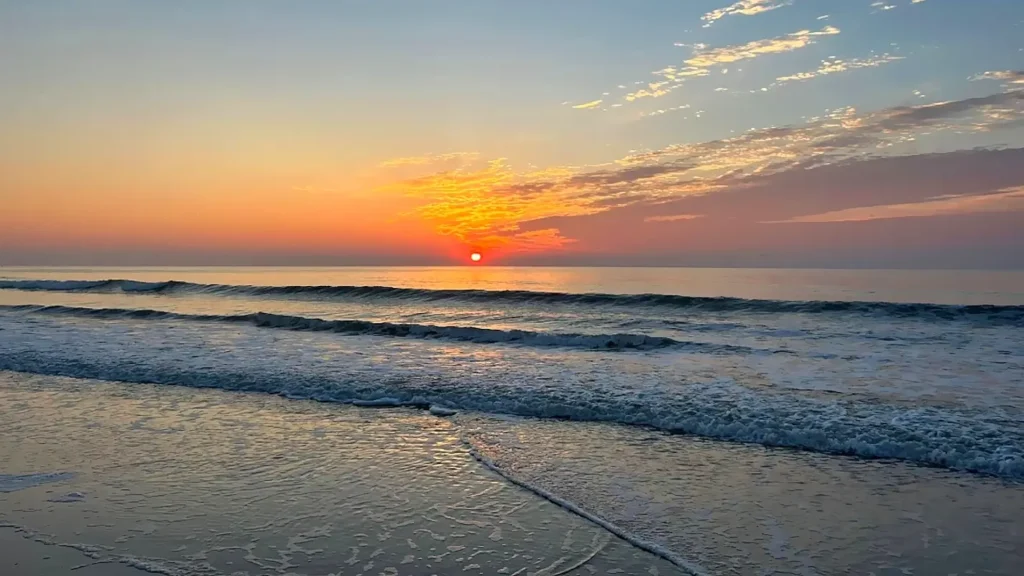 Stunning sunrise over the ocean with a vibrant orange and pink sky, the sun rising on the horizon while gentle waves roll onto the wet sand, creating reflections on the shoreline.