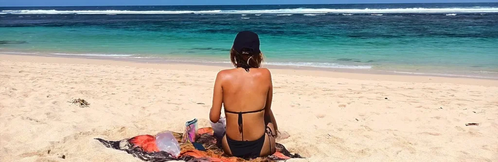 Woman sitting alone on a beach towel facing the turquoise ocean