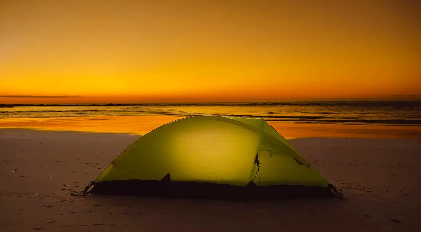 A brightly lit yellow dome tent glowing from within on a sandy beach at sunset, with a dramatic orange and yellow sky reflecting on the ocean waves in the background.
