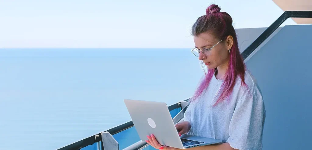 woman with pink-tipped hair working on laptop on a balcony overlooking calm water