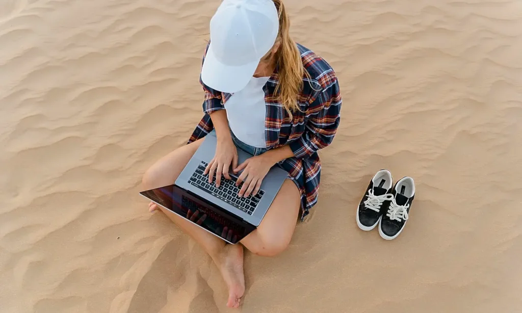 woman in white cap and plaid shirt working on laptop while sitting in sand, shoes beside her