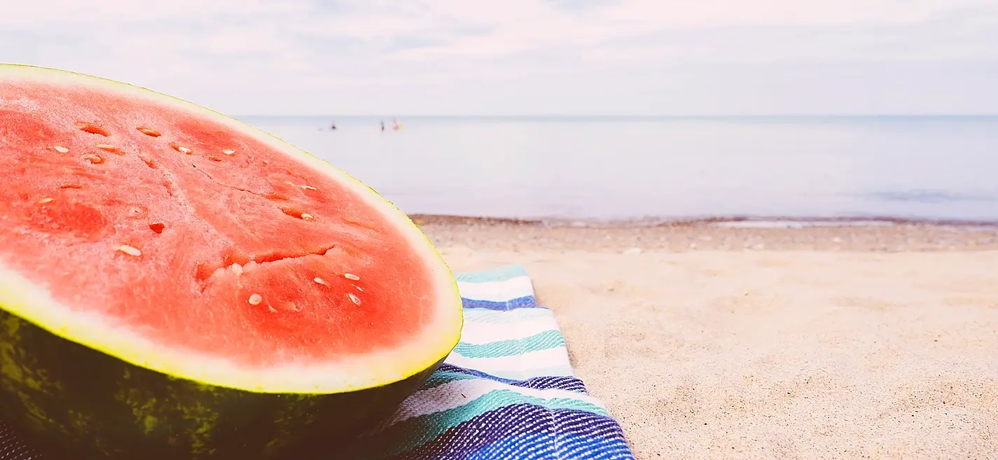 Large half watermelon resting on a striped towel on a sandy beach with the sea in the background