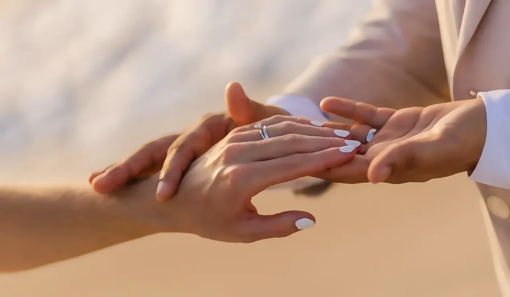 A close-up of a couple’s hands gently holding each other on the beach. The woman’s hand, with white nail polish and a sparkling diamond engagement ring, rests on the man’s hand. Warm golden sunlight illuminates the tender moment.
