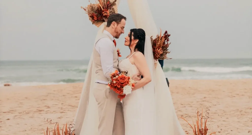A bride and groom standing close together under a wedding arch on the beach. They are gazing lovingly at each other. The groom wears a vest and the bride holds a vibrant orange and white floral bouquet. The ocean is visible in the background.