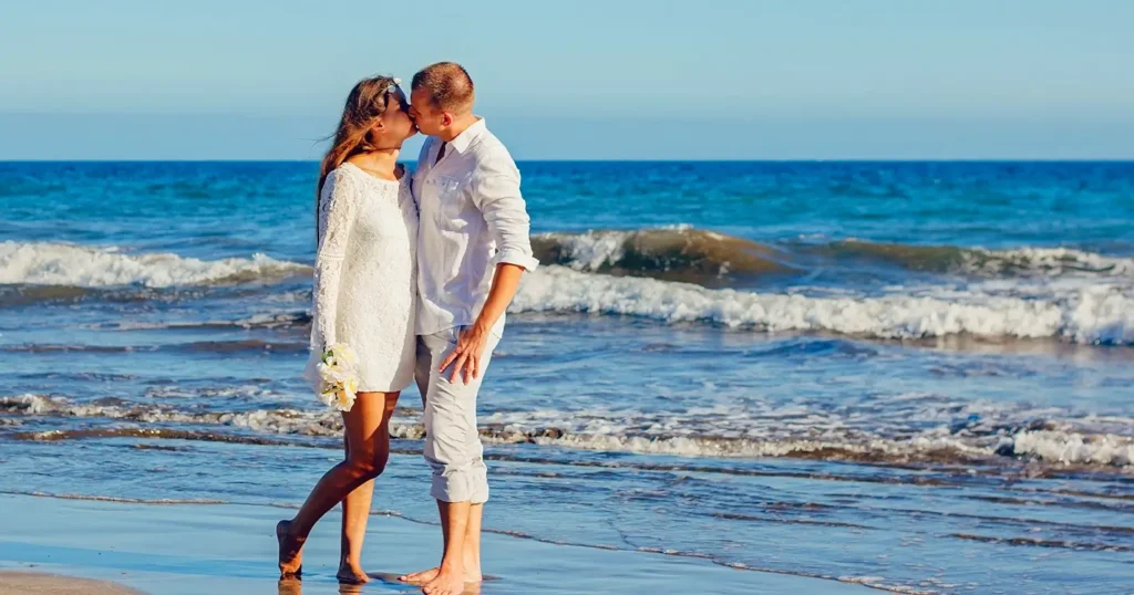 A bride and groom sharing a kiss at the water’s edge on the beach. The bride wears a white lace dress and holds a small bouquet while the groom is in a white shirt with rolled-up pants. Ocean waves gently surround their bare feet