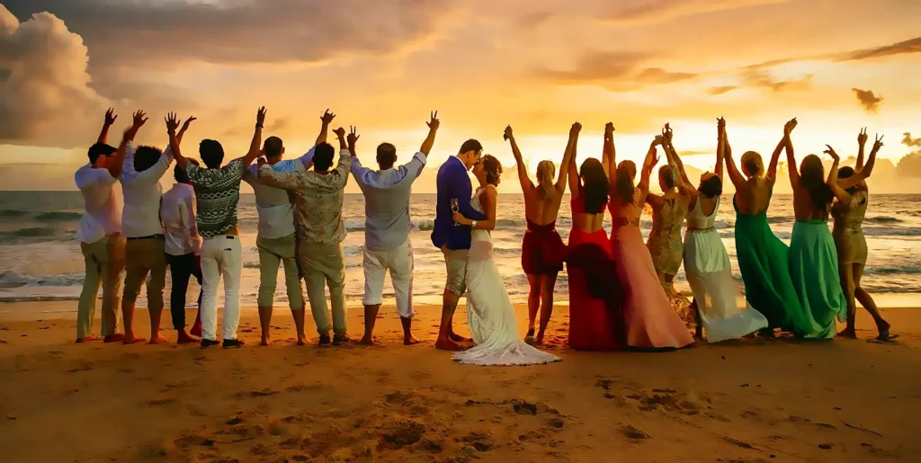 A joyful group of wedding guests and the bride and groom standing on the beach at sunset. Everyone is facing the ocean with their arms raised in celebration. The bride and groom are kissing in the center while the guests cheer around them under a golden sky.