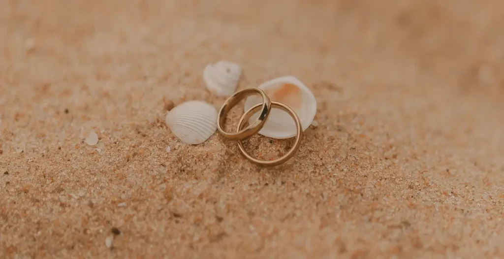 Two gold wedding rings resting on soft beach sand surrounded by small white seashells. A close-up romantic detail shot.