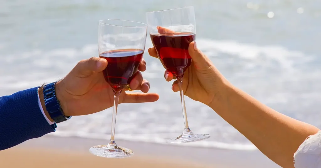 Two hands toasting with glasses of red wine on a sunny beach. One hand belongs to a person in a blue suit, the other to a person in a white dress. The ocean waves are visible in the soft-focus background.