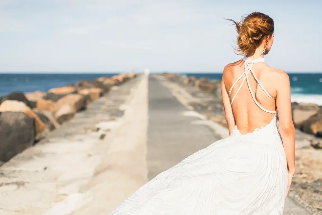 A bride viewed from behind, standing on a stone pier by the ocean. She is wearing a beautiful white backless wedding dress with intricate crisscross straps. Her hair is styled in an updo with strands blowing in the wind.
