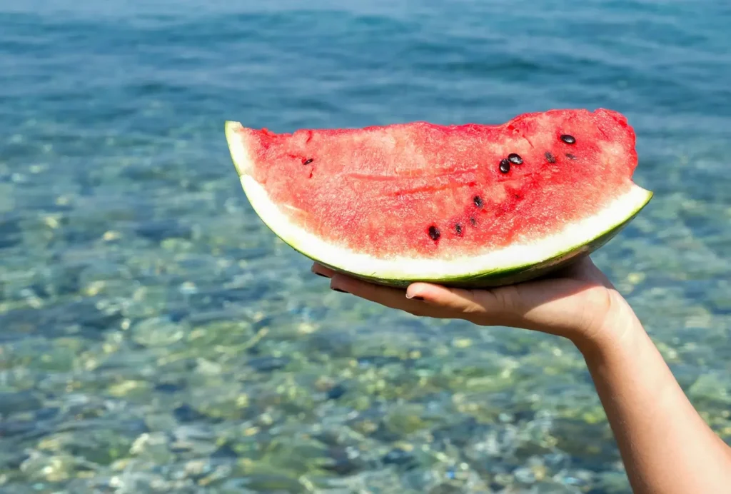 Hand holding a fresh watermelon slice over clear turquoise ocean water