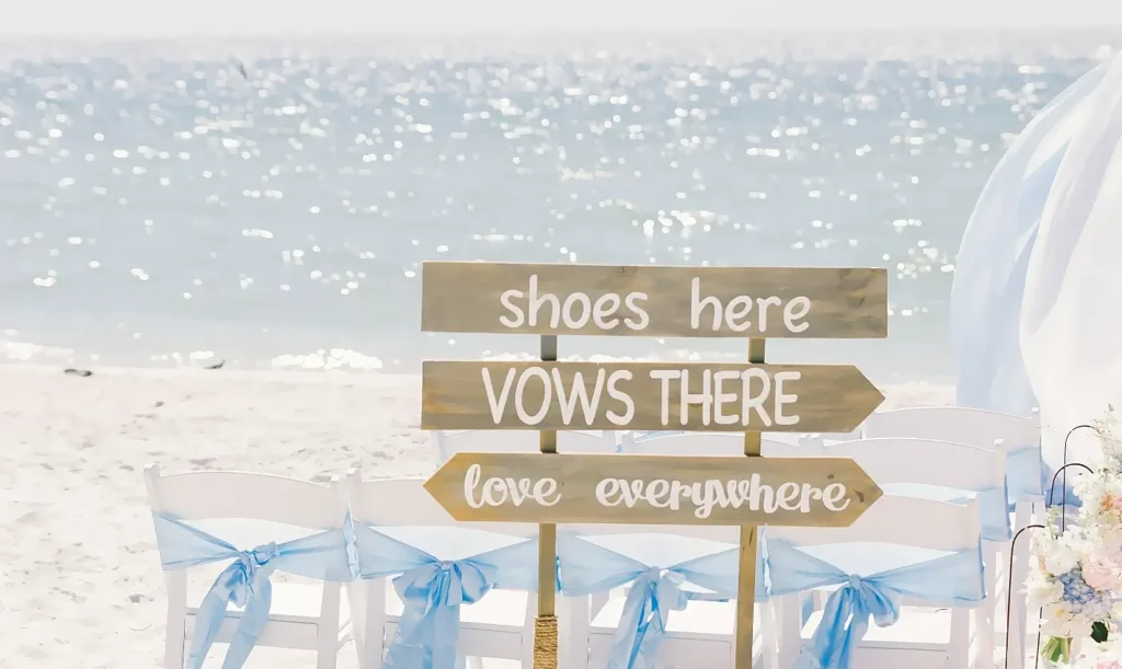 A beach wedding setup with a wooden directional signpost on the sand. The signs read “shoes here,” “VOWS THERE” with a right arrow, and “love everywhere” with a right arrow. White folding chairs decorated with light blue ribbons are arranged in front of the sign, with the ocean sparkling in the background.