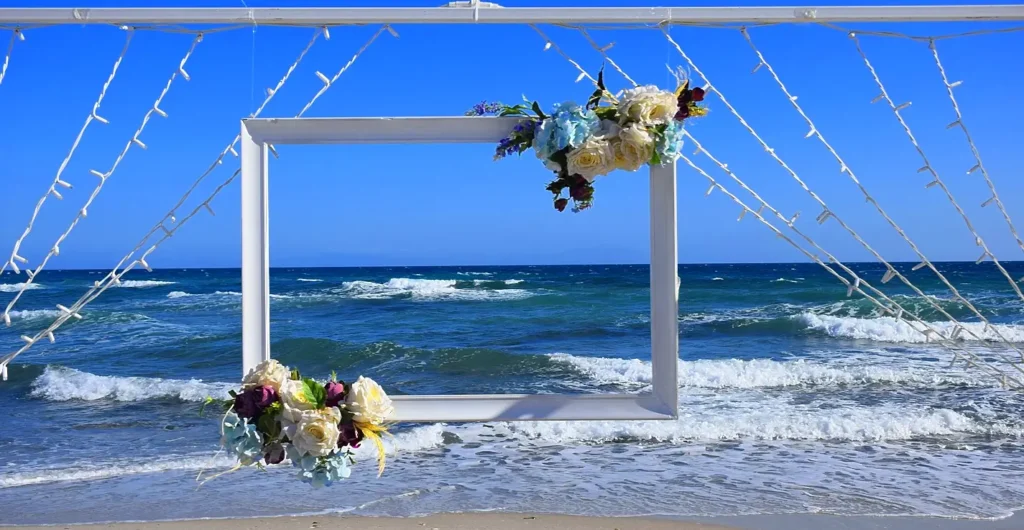 White picture frame photo booth decorated with flowers on the beach shoreline