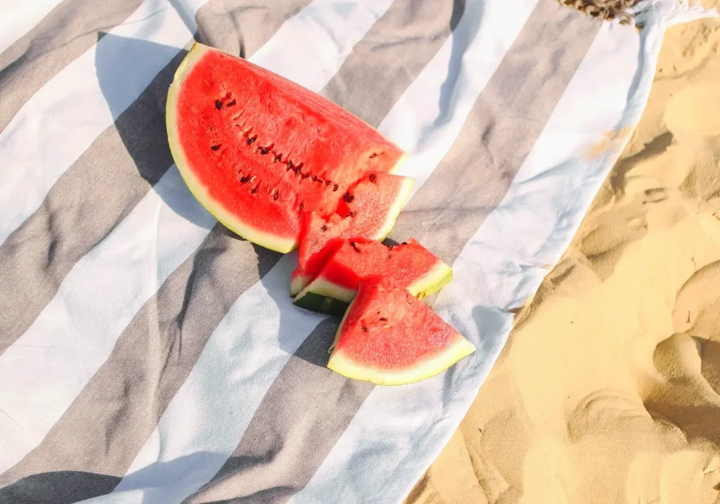 Large watermelon slice and smaller pieces resting on a striped blanket on the sand at the beach