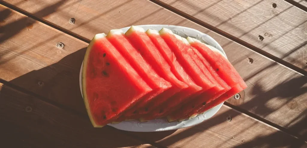 Sliced watermelon arranged on a white plate on a sunny wooden table with strong shadows