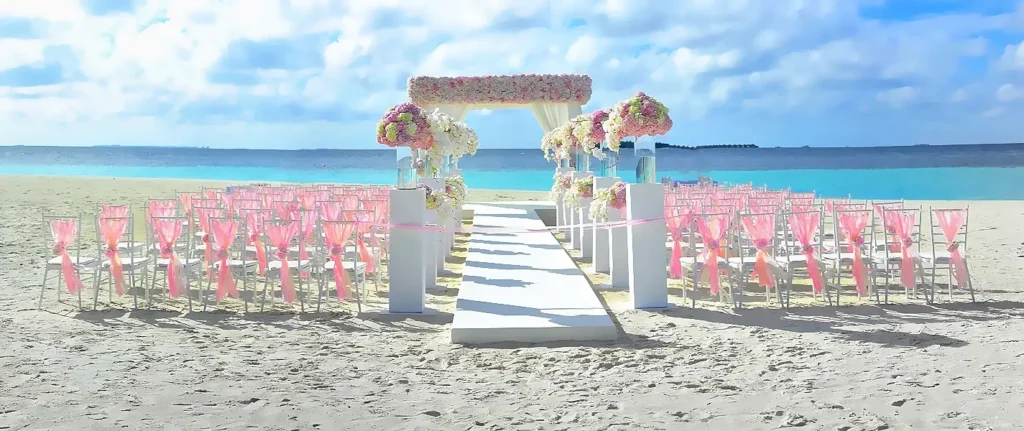 A stunning beach wedding ceremony setup with rows of white chairs decorated with pink ribbons, a white aisle runner, and a large floral arch at the front. The turquoise ocean and blue sky create a perfect tropical backdrop.