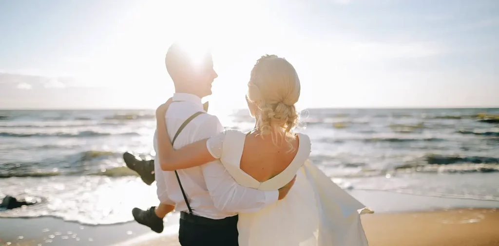 A groom in a white shirt and suspenders carrying his bride on his back on the beach. The couple is silhouetted against a bright sun with ocean waves in the background. The bride wears a white dress and has her hair in an updo.