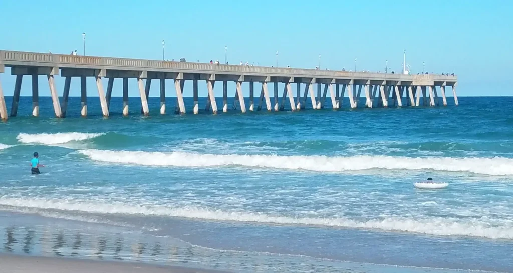 Long concrete pier extending into the blue ocean at Wrightsville Beach, North Carolina, with waves breaking in the foreground and people swimming and walking on the pier.