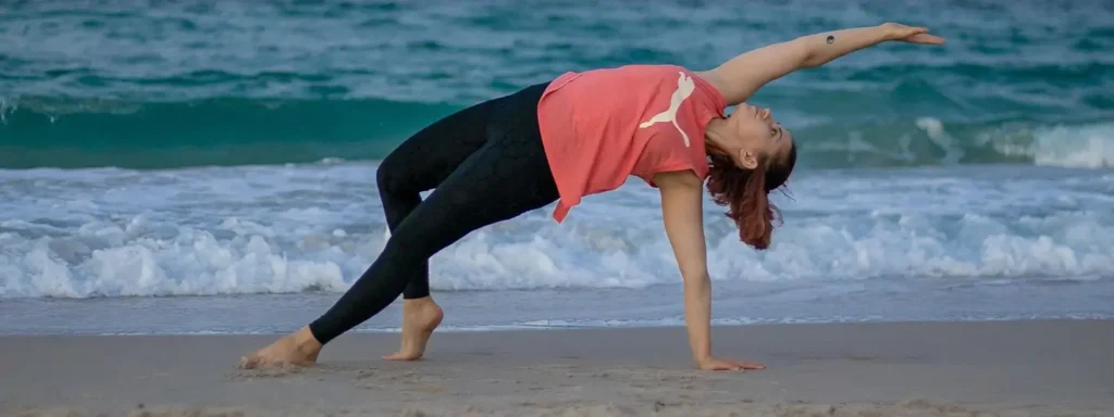 A woman in a coral t-shirt and black leggings performing a side stretch yoga pose on the beach, one hand on the sand and the other extended upward, with ocean waves behind her.