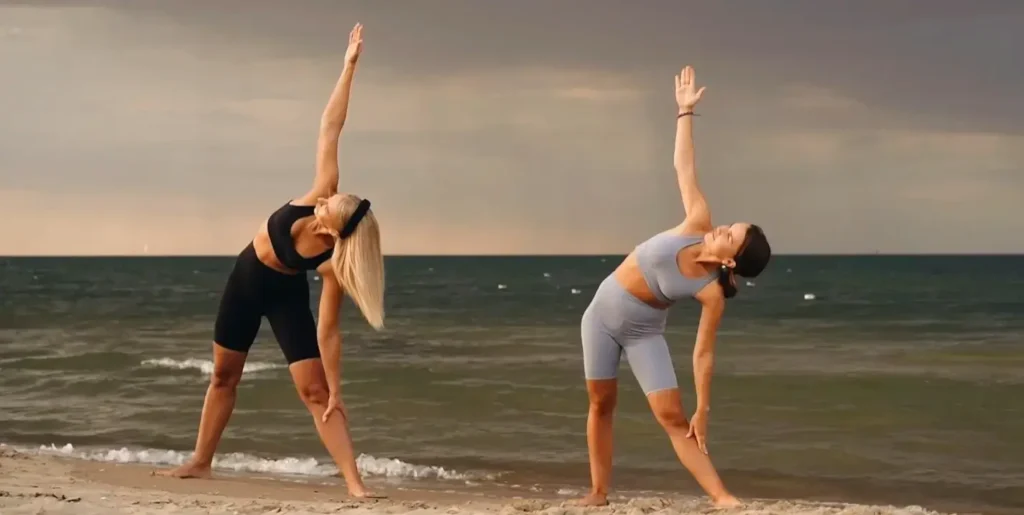 Two women practicing triangle pose side-by-side on the beach, both with one arm extended upward and the other reaching toward their leg, ocean and overcast sky in the background.