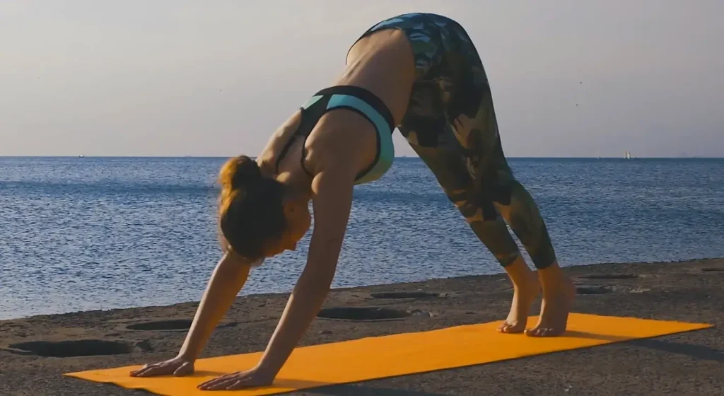 A woman in camouflage-patterned leggings and a sports bra performing downward-facing dog on a bright yellow yoga mat on a rocky coastal area overlooking the sea.