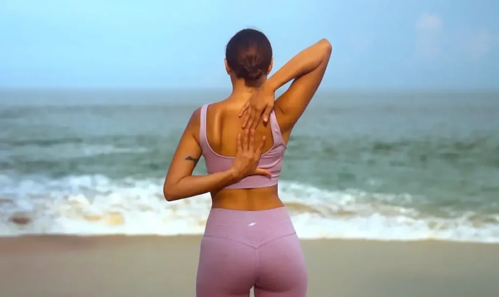 Rear view of a woman in a light pink athletic outfit standing on the beach, performing a shoulder stretch with one hand behind her head and the other on her upper back, facing the ocean.