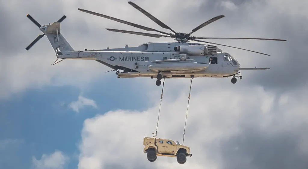 A U.S. Marine Corps CH-53E Super Stallion heavy-lift helicopter in flight, carrying a tan military Humvee-style vehicle suspended by cables beneath it against a blue sky with scattered clouds.