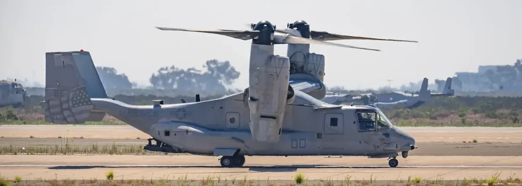 A U.S. Marine Corps MV-22 Osprey tiltrotor aircraft parked on an airfield runway, with its proprotors tilted upward in helicopter mode and an American flag design on the tail.