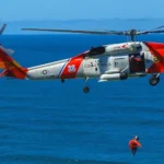 A U.S. Coast Guard MH-60 Jayhawk helicopter in bright orange and white livery hovering over the ocean, with a rescuer in an orange suit being lowered by hoist toward the water.