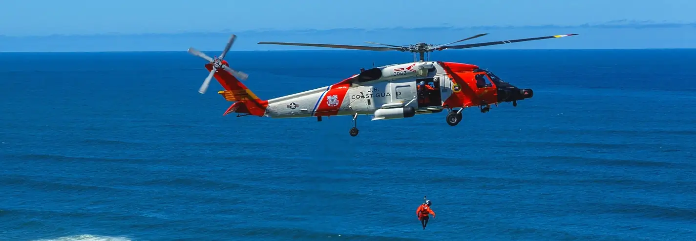 A U.S. Coast Guard MH-60 Jayhawk helicopter in bright orange and white livery hovering over the ocean, with a rescuer in an orange suit being lowered by hoist toward the water.