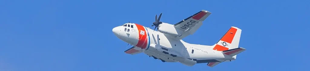 A U.S. Coast Guard HC-130 Hercules aircraft in flight against a clear blue sky, showing its distinctive white, orange, and blue color scheme with USCG markings.