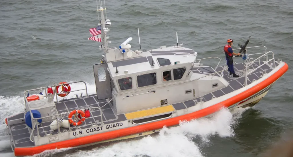 A U.S. Coast Guard response boat speeding through choppy water, creating a large wake, with a crew member in tactical gear holding a rifle on the rear deck.