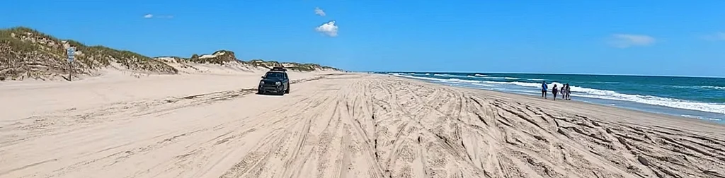 A 4x4 SUV driving the sand beach in Corolla's 4WD area while beachgoers walk along the shoreline