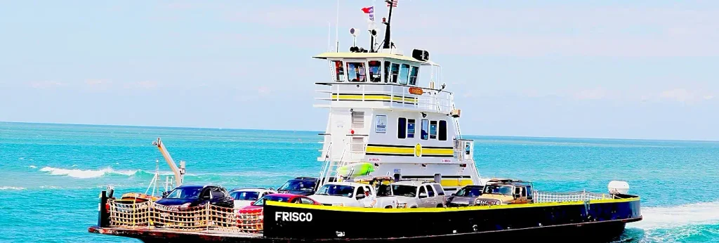 North Carolina Ferry System vessel named FRISCO carrying cars across turquoise water under a bright blue sky, with American flags flying and passengers visible on the upper deck – Hatteras–Ocracoke ferry.