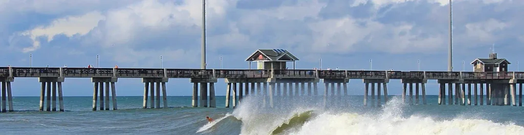 Jennette’s Pier in Nags Head stretching into the Atlantic under partly cloudy skies, with people walking the pier and a surfer riding a breaking wave in the foreground.