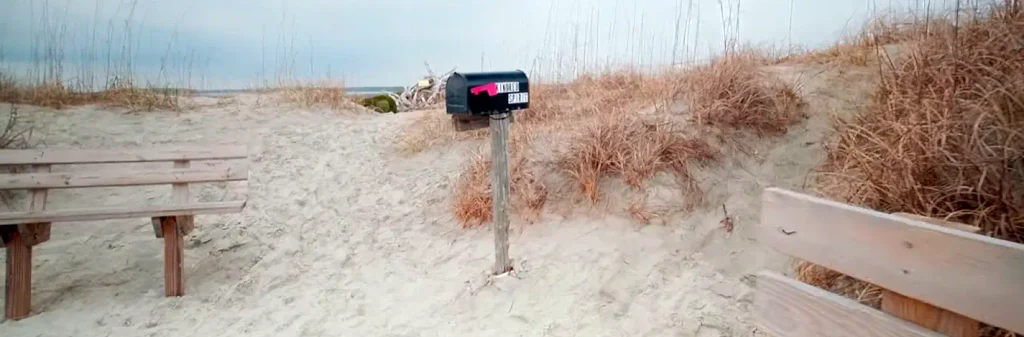 Black “KINDRED SPIRIT” mailbox with pink North Carolina shape centered on a sandy path between two wooden benches, surrounded by dry dune grass with the ocean visible in the distance under gray skies.