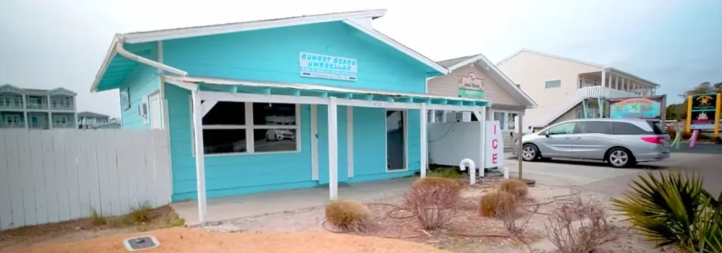 Bright turquoise beach shop labeled “Sunset Beach Umbrellas” with white trim, an ice machine, parked vehicles, and nearby beach houses under an overcast sky.