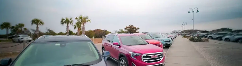 Busy paved parking area filled with cars and SUVs, palm trees, streetlights, and beach access path under a cloudy sky near Sunset Beach, North Carolina.