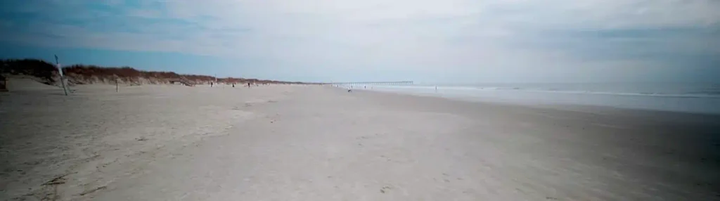 Expansive empty stretch of light sand beach leading to the ocean and a distant pier, with low dunes and a few distant people under an overcast sky at Sunset Beach, NC.