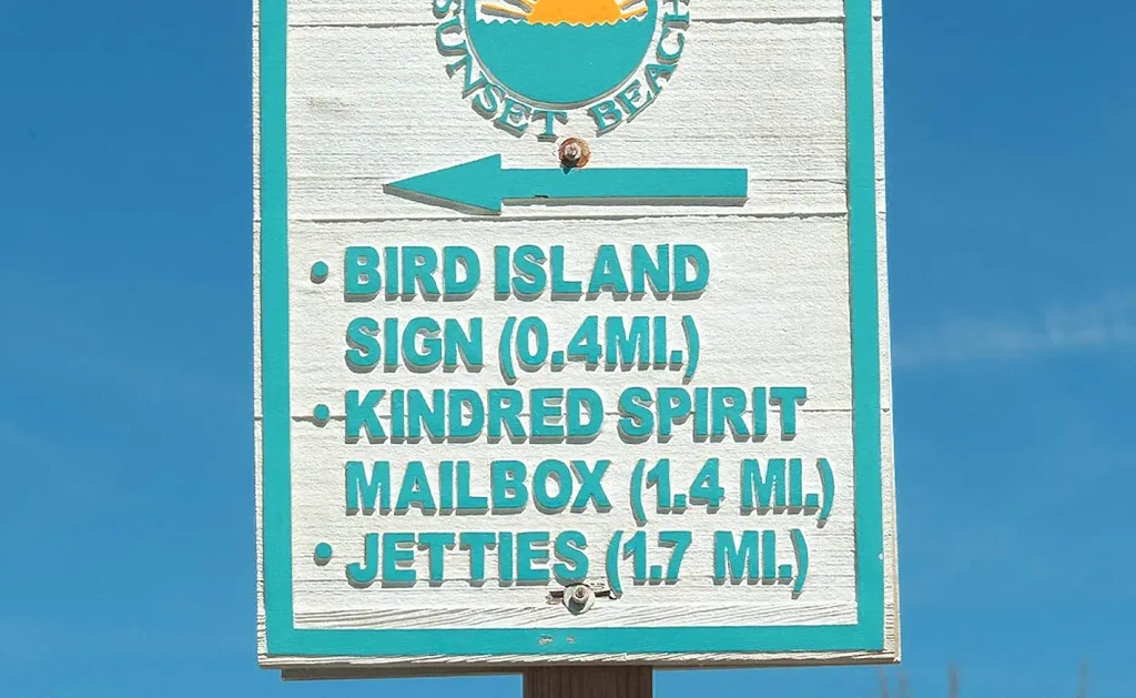 White wooden sign with teal lettering and a Sunset Beach sun logo pointing left toward Bird Island Sign (0.4 mi.), Kindred Spirit Mailbox (1.4 mi.), and Jetties (1.7 mi.) under a blue sky.