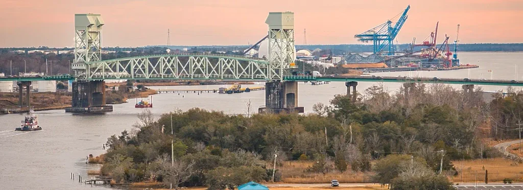 Green vertical-lift bridge spanning a river at sunset, with industrial cranes and port facilities in the background and a small tugboat on the water.