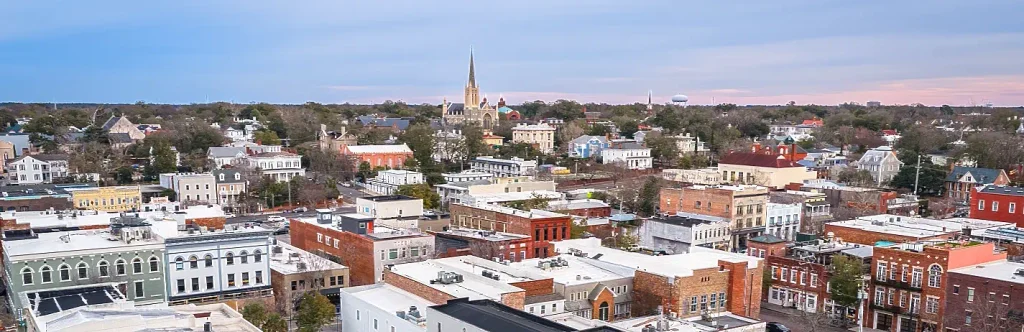 Panoramic rooftop view of a historic downtown with low brick and painted buildings, a prominent church steeple rising in the distance, and soft evening light.