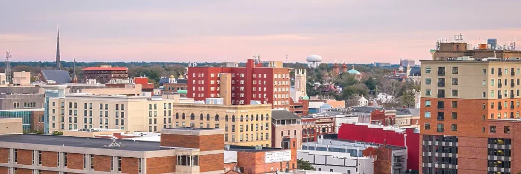 Elevated view of a mid-sized city’s skyline with red-brick and beige buildings, a tall red hotel, church steeples, and a distant water tower under a pink-tinged sky.