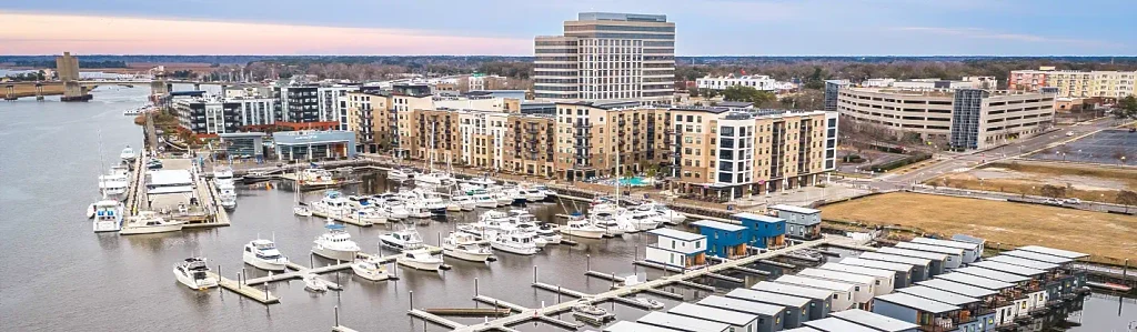 Aerial view of a busy marina filled with white boats and yachts, lined by modern multi-story residential and commercial buildings along the waterfront at dusk.