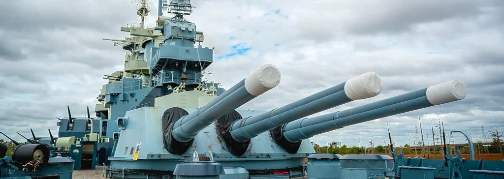 Close-up of the massive triple-gun turrets of the battleship USS North Carolina, with white muzzle covers, on a cloudy day.