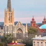 Historic gray stone Gothic Revival church with tall beige spire and pointed arches, next to a red brick building with twin towers and a turquoise dome, surrounded by trees and houses under a soft pastel sky.
