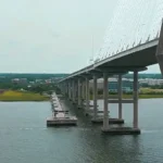 Wide view of the Arthur Ravenel Jr. Bridge (cable-stayed span) and the parallel Mount Pleasant Pier stretching into Charleston Harbor, with green shoreline, buildings, and a water tower in the distance under overcast skies.