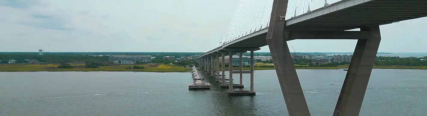 Wide view of the Arthur Ravenel Jr. Bridge (cable-stayed span) and the parallel Mount Pleasant Pier stretching into Charleston Harbor, with green shoreline, buildings, and a water tower in the distance under overcast skies.